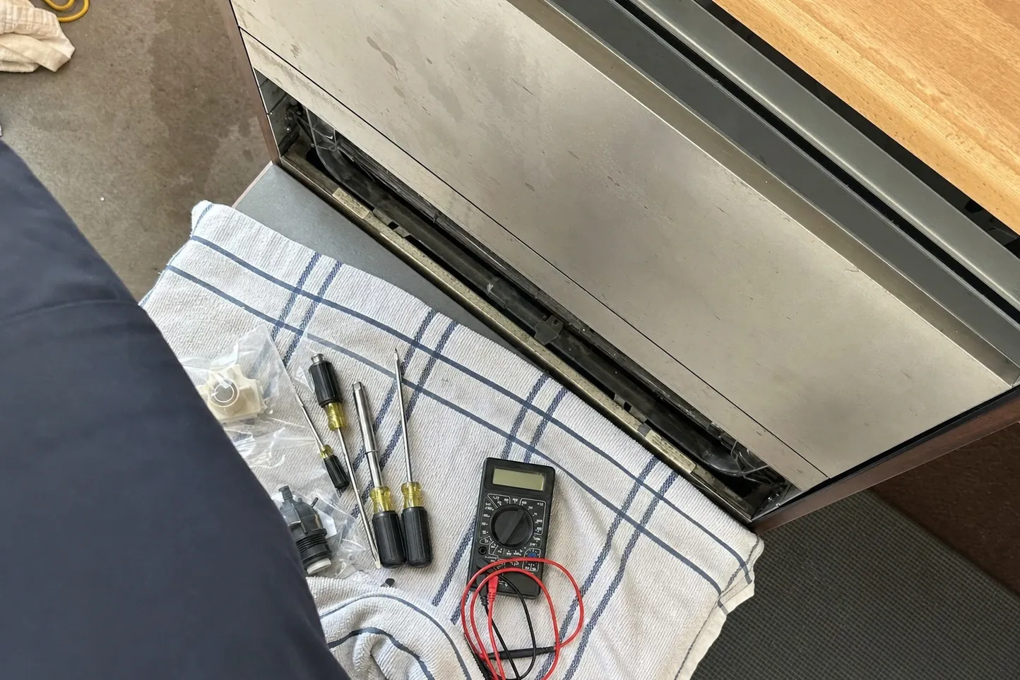 Multimeter, screwdrivers and two bagged replacement parts laid out on a folded kitchen towel beside a stainless undercounter unit opened a few inches